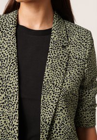 Grüner Blazer mit schwarzem Leopardenmuster, ausgestattet mit einem Reverskragen und aufgerollten Ärmeln, getragen über einem schwarzen Oberteil. Glatte Stofftextur.