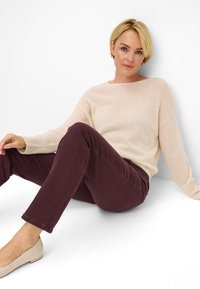 Beige, hyggelig trøje med løs pasform, parret med vinfarvede slim pants. Modellen sidder imod en hvid baggrund iført lyse sko.
