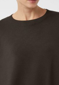 Zbliżenie na osobę w ciemnoszarym swetrze z prążkowanym dekoltem typu crew neck, ukazujące linię dekoltu i górną część klatki piersiowej.