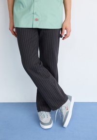 Cămașa verde mentă cu nasturi, pantaloni negri cu dungi subțiri și adidași gri cu accente roșii pe o suprafață albastră. Ținuta este simplă și casual.