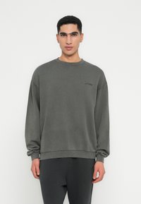 Přehnaný crewneck svetr v tmavě šedé barvě, vyrobený z bavlněné směsi. Obsahuje žebrované manžety a lem, s malým logem na hrudi.
