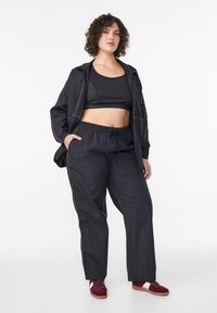 Schwarzes Sportoutfit bestehend aus einem bauchfreien Tanktop, lockeren Hosen mit elastischem Bund und einer schwarzen Zip-up-Jacke mit pinkfarbenen Akzenten.