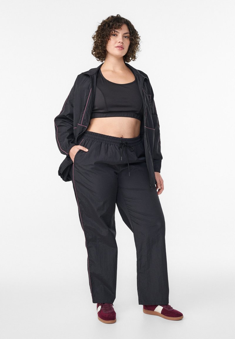 Schwarzes Sportoutfit bestehend aus einem bauchfreien Tanktop, lockeren Hosen mit elastischem Bund und einer schwarzen Zip-up-Jacke mit pinkfarbenen Akzenten.