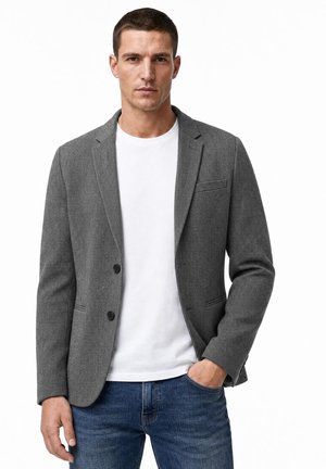 Man met een grijze blazer, wit T-shirt en blauwe jeans, staand met één hand in zijn zak tegen een witte achtergrond.