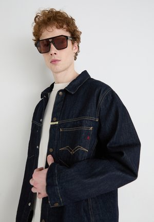 Denim jacket in dark blue with gold stitching, button closure, and two chest pockets. Model wearing oversized sunglasses and a white shirt.