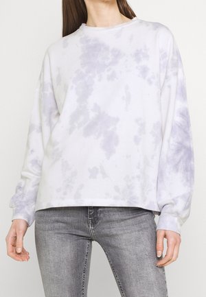 Kobieta ubrana w luźną białą i jasnofioletową bluzę tie-dye oraz dopasowane szare dżinsy, stoi z rozluźnionymi ramionami.