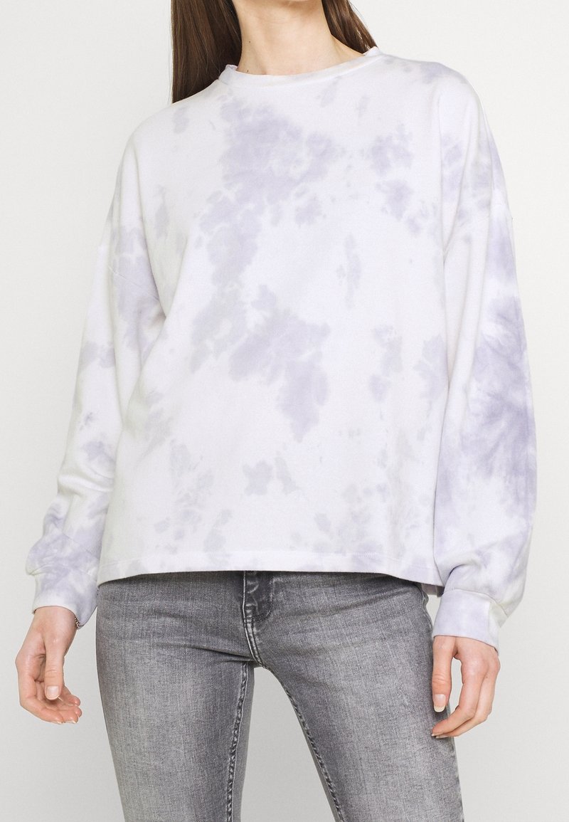 Femme portant un sweat-shirt ample blanc et violet clair tie-dye et un jean gris ajusté, debout avec les bras détendus.