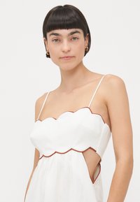 White dress with a scalloped neckline and brown trim, featuring thin straps and cut-out sides. Smooth fabric with a flowing design.