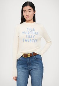 Valge kootud kampsun helesinise tekstiga "KÜLM ILM MUGAV KAMPSUN." Soonikkoes varrukaotsad ja ääred. Modell kannab siniseid teksapüüre ja pruuni vööd.