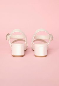 Sandalias de satén blanco con los dedos al aire y un tacón grueso, que cuentan con correas en el tobillo traseras y un acento circular decorativo en el tacón.