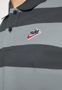 Detailní záběr na šedý pruhovaný polo tričko s černými knoflíky a červeným a černým logem Nike na hrudi.