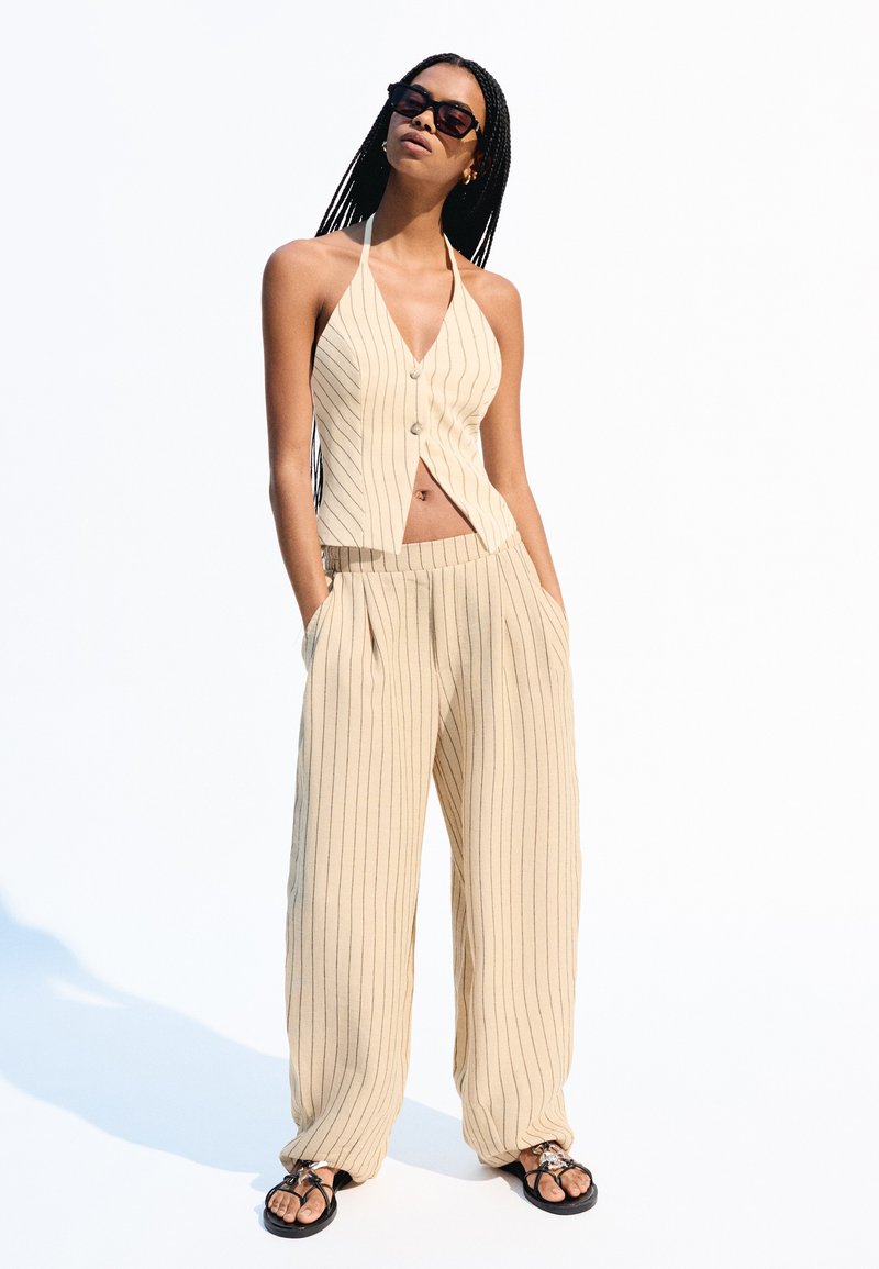 Beige gestreifter Neckholder-Top mit Knopfverschluss, kombiniert mit hoch taillierten, weiten Hosen. Schwarze Sandalen runden den Look ab, vor einem weißen Hintergrund.