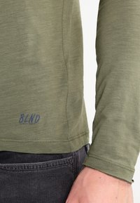 Long-sleeve shirt in olive green, featuring a textured fabric, stitched hem, and a visible "B.L.N.D" logo in navy on the lower side.