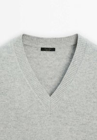 Pull en maille gris clair à col en V avec col côtelé et une étiquette de marque noire cousue à l’intérieur de l’encolure.