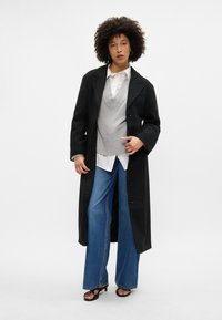 Dlouhý černý kabát, šedý svetr s žebrovanou strukturou, bílá košile, modré široké džíny, černé sandály na pásky, jednobarevné pozadí.