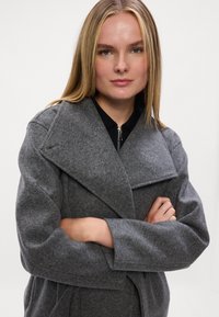 Siv volnen plašč iz mešanice volne s širokim ovratnikom, dolgimi rokavi in podrobnostmi na reverju. Model z crossed arms, črna notranja plast vidna.