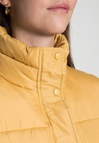 Gelbe gepolsterte Jacke mit einem hohen Kragen, die sichtbare Nähte und gelbe Druckknöpfe aufweist. Glattes, synthetisches Material mit einer weichen Textur.