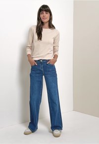 Vaaleanbeige pitkähihaisessa paidassa on kireät hihat, ja se on yhdistetty leveälahkeisiin sinisiin farkkuihin. Malli seisoo kädet taskuissa ja hänellä on päällään rentoja kenkiä.