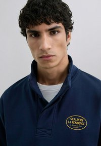 Sudadera azul marina con un alto cuello, que presenta un logo bordado en amarillo que dice "SCALPERS LA RÉSIDENCE, PARÍS." Textura suave.