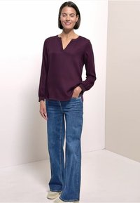 Dieprode blouse met lange mouwen, een V-hals en knopen, gecombineerd met blauwe wijde jeans en lichtgekleurde schoenen, tentoongesteld tegen een neutrale achtergrond.