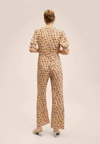 Tuta floreale con maniche a sbuffo corte, sfondo beige con fiori arancioni e bianchi, vita aderente e design a gamba larga.