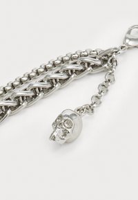 Bracelet en chaîne argentée avec des maillons entrelacés et un trésor en forme de crâne. Le trésor présente des traits faciaux détaillés et une texture lisse.