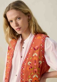 Gilet brodé orange avec des motifs floraux, featuring des accents roses, verts et blancs, sur une chemise à boutons rose clair. Texture lisse.