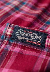 Tissu à carreaux rouge et bleu avec une texture douce ; présente une étiquette noire avec "SuperDry" et "Shirting Co." brodés en argent et orange.