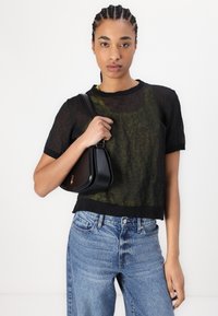Black sheer top with short sleeves over a green inner layer, paired with light blue distressed jeans. Holds a small black handbag.