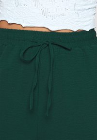 Groene broeken met een elastische tailleband en een strikkoord. De stof lijkt zacht met een subtiele textuur, wat de ontspannen pasvorm benadrukt.