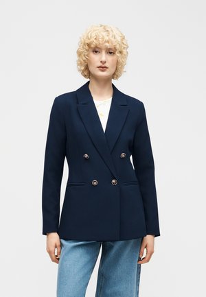 Blond krøllet person iført en marineblå dobbeltradet blazer over en hvid top med lyseblå jeans, stående mod en ensfarvet baggrund.