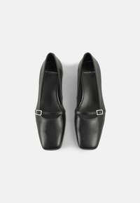 Chaussures en cuir noir avec un design à bout carré, dotées d'une fine sangle avec une boucle argentée rectangulaire. Texture lisse et style minimaliste.
