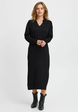 Black long-sleeve dress with a V-neck and collar, made of a soft material. Features a side slit and falls below the knee, paired with black boots.