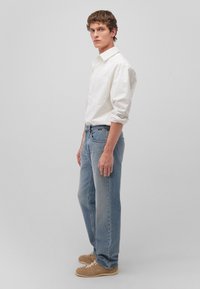 White button-up shirt, light blue jeans, and beige sneakers. The jeans have a relaxed fit, and the shirt features a single chest pocket.