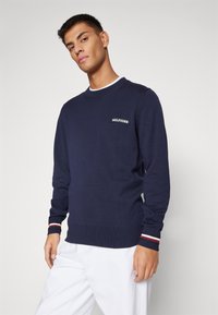 Maglione lavorato a maglia blu navy con scollatura rotonda, polsini a costine con strisce rosse e bianche, e un piccolo logo "HILFIGER" sul petto.