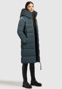 Cappotto lungo imbottito in verde scuro, con colletto alto, design trapuntato, due zip laterali e vestibilità dritta, abbinato a stivali neri fino al ginocchio.