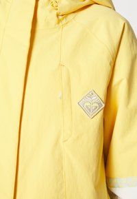 Gelbe Außenjacke mit Kapuze, frontalem Reißverschluss und einem rautenförmigen gestickten Logo auf der linken Brust. Glattes Gewebe mit genähten Akzenten.