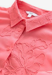 Camicia rosa corallo con bottoni, con ricami floreali sul colletto. Presenta piccoli bottoni abbinati e un tessuto morbido e strutturato.