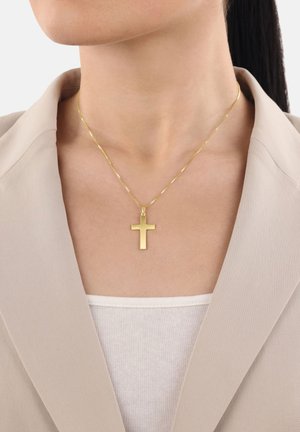 Złoty naszyjnik z prostym wisiorkiem w kształcie krzyża, posiadający skręcony łańcuszek i minimalistyczny design, noszony na białej bluzce i beżowej marynarce.