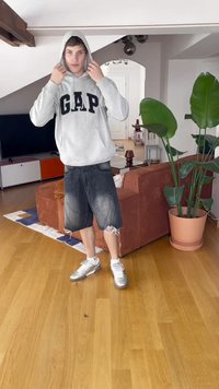 Grijze GAP hoodie, zwarte denim shorts met rafelige randen, en zilveren sneakers. De kamer heeft een bruine bank en een grote groene plant.