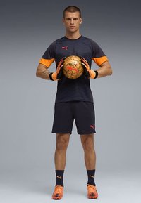 Sportaš u crno-narančastoj Puma nogometnoj opremi drži zlatnu nogometnu loptu s Puma logotipom, stoji protiv jednostavne sive pozadine.