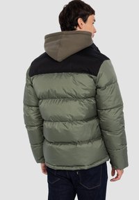 Zielona kurtka puchowa z czarną górną częścią, wyposażona w kaptur. Materiał wydaje się błyszczący, pikowany z wyraźnie widocznymi przeszyciami.