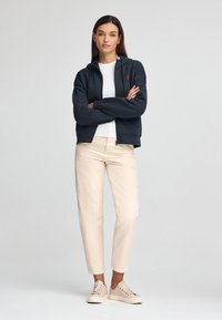 Felpa grigia scura con zip e cappuccio, t-shirt bianca sotto, pantaloni beige su misura e sneakers chiare, in piedi contro uno sfondo neutro.