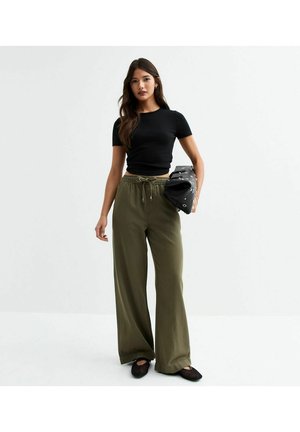 Zwarte getailleerde t-shirt, olijfgroene wijde broek, gestructureerde stof, trekkoord in de taille, en een zwarte clutch tas met zilveren accenten.