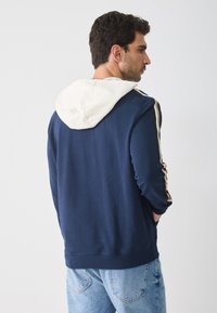 Felpa con cappuccio blu navy e interno della cappuccio bianco, caratterizzata da strisce dorate lungo le maniche. Materiale in cotone morbido, orlo e polsini a costine.
