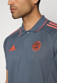 Grå polo shirt med en tekstureret finish, orange detaljer på kraven og ærmerne, samt et broderet FC Bayern München-logo på brystet.