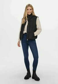 Svart pufferväst med hög krage, dragkedjeförslutning, i kombination med en beige tröja, blå jeans och svarta ankelstövlar. Slät textur, avslappnad passform.