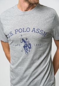 Graues Baumwoll-T-Shirt mit Rundhalsausschnitt, das ein blaues Grafikdesign mit einem Polo-Spieler zeigt, und dem Text "U.S. POLO ASSN" darüber sowie "SINCE 1890" darunter.
