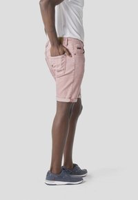 Homme portant un short en jean rose clair retroussé, un t-shirt blanc et des chaussures décontractées bleu marine, debout avec une main dans la poche arrière.