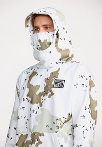 Sudadera con capucha blanca que presenta un patrón de camuflaje en tonos beige y oscuros, capucha ajustable y parche con logo en el frente. Textura de tela suave.